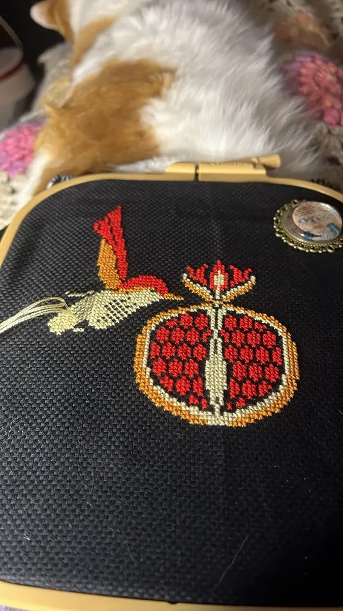 Buyer photo of the Pomegranate Bird design in progress on black fabric in a wooden hoop, with the red and gold pomegranate finished and a cat napping behind